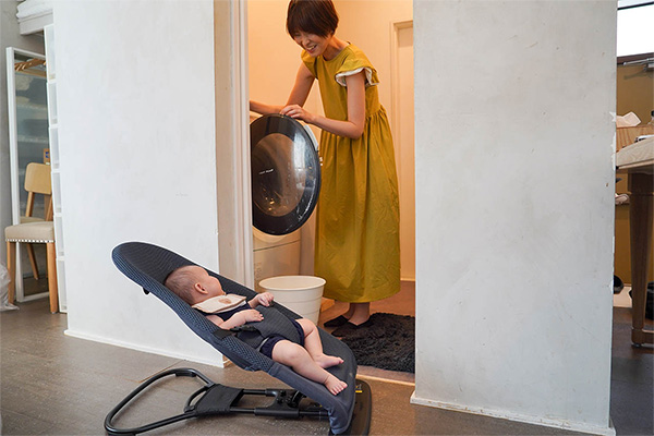 洗濯機の蓋をあけながら、近くに置いたバウンサーの上の赤ちゃんを笑顔で見ている女性