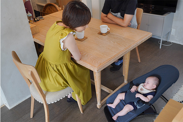 ダイニングテーブルに座った大人二人と、近くに置いたバウンサーの上の寝ている赤ちゃん